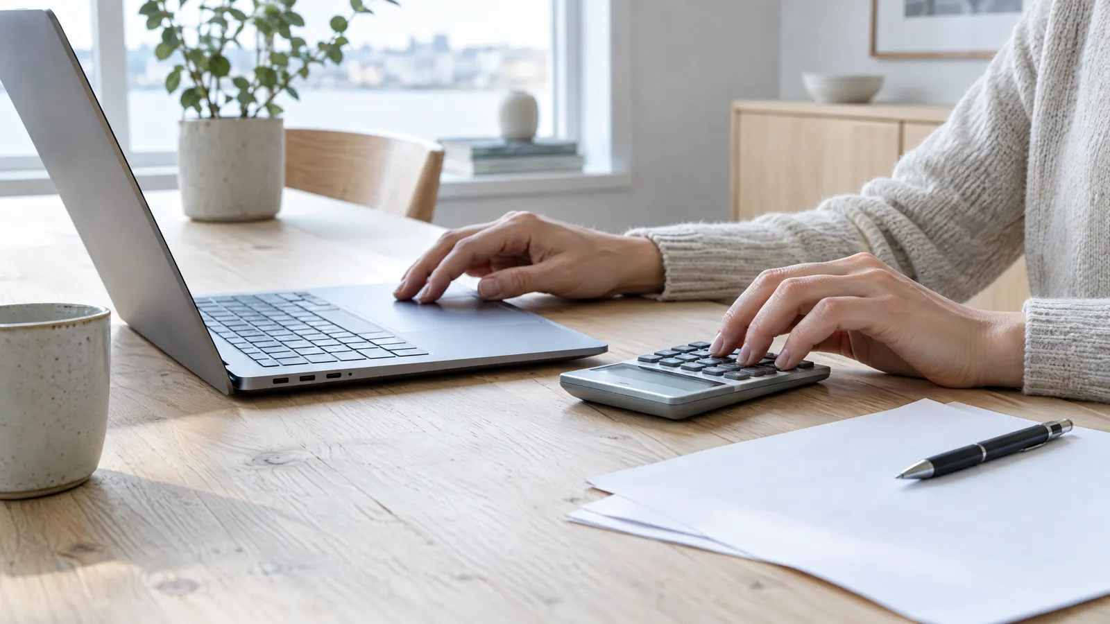 Hands at a laptop and a calculator on a wooden kitchen table, with paper and a pen beside them